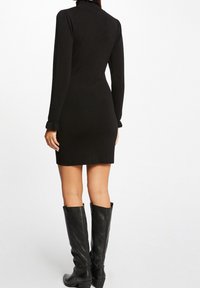 Robe noire à manches longues avec un col haut, silhouette ajustée et poignets à volants, accompagnée de hautes bottes en cuir noir.