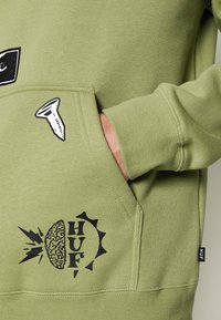 Felpa verde con tasca frontale; presenta design stampati in nero tra cui una vite, un'immagine di un cervello e la parola "HUF". Texture morbida.