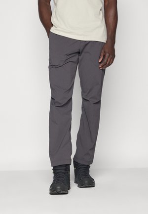 Homme portant un pantalon gris foncé, une chemise beige clair à manches courtes et des chaussures de randonnée noires, debout devant un fond blanc.