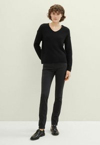 Juodas megztinis su V formos iškirpte, derinamas su juodomis lieknomis kelnėmis ir juodais batais. Megztinis turi tekstūruotą raštą.