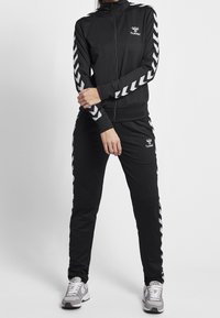 Svart träningsoverall med en zip-up jacka och matchande byxor, som har vita chevron-accenter på ärmarna och sidorna. Tyget har en mjuk textur.