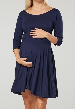 Kobieta w ciąży, ubrana w granatową sukienkę, trzymająca obie ręce na brzuchu, stojąca na tle jednolitego jasnego tła.