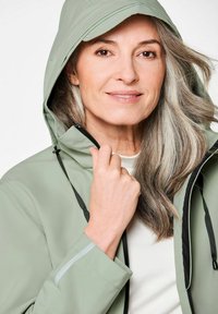 Lichtgroene waterdichte jas met een capuchon, rits aan de voorkant en verstelbare manchetten. Heeft zwarte accenten en een gladde textuur.