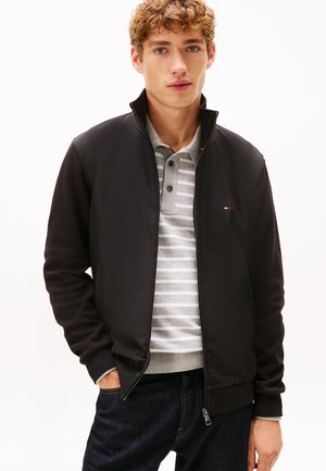 Chaqueta negra con cremallera y textura suave, con cuello alto, combinada con una camiseta polo gris de rayas y vaqueros oscuros.