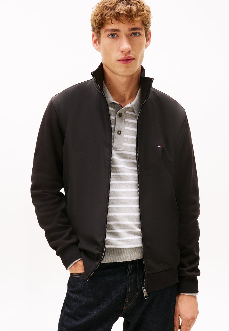 Sort lynlåsjakke med en glat tekstur, med en ståkrave, kombineret med en gråstribet poloskjorte og mørke jeans.