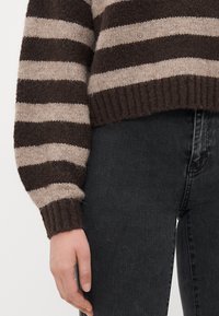 Hand resting by side wearing a brown and beige striped knit sweater with dark gray jeans.
