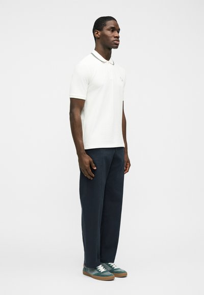 Άνδρας στέκεται πλευρικά, φοράει λευκή κοντομάνικη πόλο, σκούρο μπλε παντελόνι και τιρκουάζ αθλητικά παπούτσια με λευκά κορδόνια σε απλό φόντο.