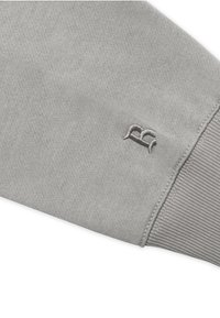 Grauer Sweatshirt mit weicher Textur, ausgestattet mit einem gerippten Bündchen und einem gestickten Logo in der Nähe des Ärmelabschlusses. Einfaches, minimalistisches Design.