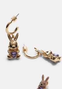 Guldtonede øreringe med en kanincharme, der holder en lilla hjerteformet ædelsten. Detaljeret design med skinnende finish og hoop-baggrund.