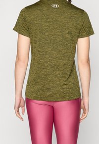 Mujer con camiseta deportiva de manga corta color verde oliva y mallas rosas brillantes, de pie con la espalda hacia la cámara.