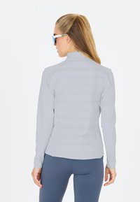 Lys grå atletisk jakke med høj krave, der har en quiltet tekstur og lange ærmer, parret med marineblå leggings og solbriller.