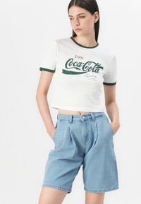 Cropped white t-shirt with green ringer detail and Coca-Cola graphic; blue high-waisted denim shorts with pleats and side pockets.