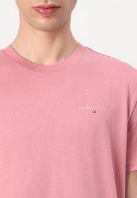 Rózsaszín pamut póló kerek nyakkivágással és rövid ujjakkal. A mellkason egy kis, fehér "TOMMY JEANS" logó található. Simaszerkezet.