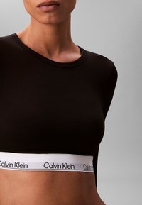 Black long-sleeve top with a round neckline and a white elastic band featuring "Calvin Klein" branding at the hem. Smooth texture.
