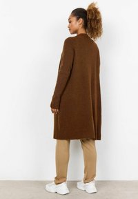 Brown knitted cardigan featuring long sleeves and a relaxed fit, paired with beige pants and white sneakers. The fabric appears soft and comfortable.
