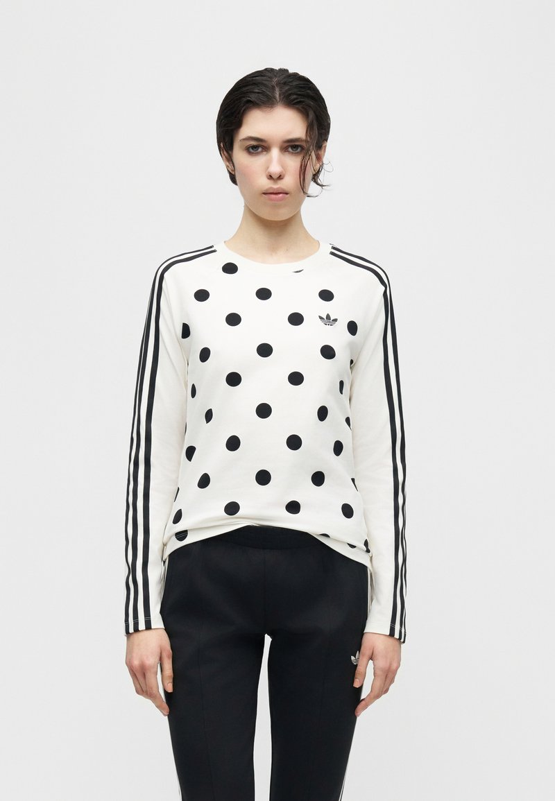 Ung person med kort mørkt hår iført hvid langærmet skjorte med sorte polkaprikker og sorte striber på ærmerne samt sorte bukser.