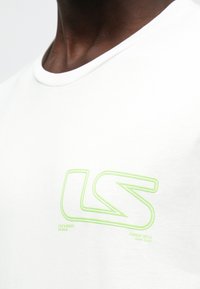 Hvit bomulls-T-skjorte med rund hals og korte ermer. Har en grønn brodert "LS"-logo på brystet. Myk tekstur.