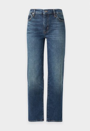 Femme portant un haut à manches longues foncé à motifs, un jean bleu et des ballerines noires pointues, debout avec une main dans la poche sur un fond uni.