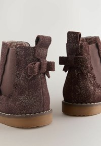 Brązowe ankle boots z brokatem o miękkiej zamszowej teksturze. Zawierają detal w postaci kokardy oraz faliste krawędzie, osadzone na gumowej podeszwie.
