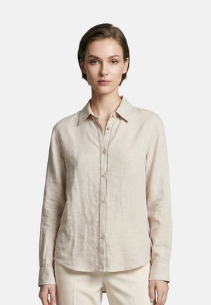 Giovane donna con capelli corti che indossa una camicia beige a maniche lunghe con bottoni e pantaloni chiari su uno sfondo semplice.