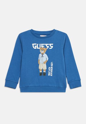 Sudadera azul de manga larga con un dibujo de un oso de peluche con pantalones cortos y camisa, texto "GUESS" arriba y texto vertical "Los Angeles EST. 1981" en el frente.