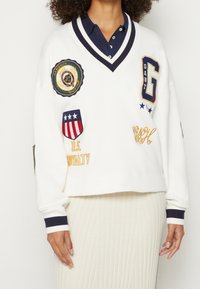 Sweat-shirt blanc court avec accents bleu marine, col en V, et plusieurs patchs brodés, comprenant un logo, des étoiles et des détails textuels.