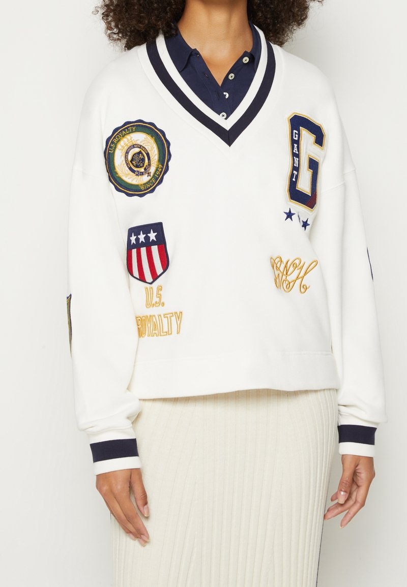 Sweat-shirt blanc court avec accents bleu marine, col en V, et plusieurs patchs brodés, comprenant un logo, des étoiles et des détails textuels.