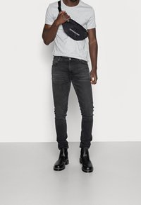 Man som bär en ljusgrå t-shirt, svarta slim fit-jeans, svarta läderstövlar och en svart Calvin Klein crossbody-väska.