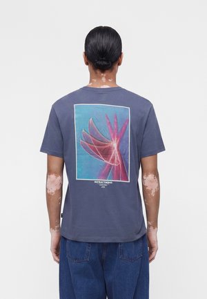 Άτομο με λεύκη και μαλλιά δεμένα πίσω φοράει ένα σκούρο μπλε πουκάμισο με πολύχρωμο αφηρημένο γραφικό ενός χεριού σε κίνηση στην πλάτη και μπλε τζιν.