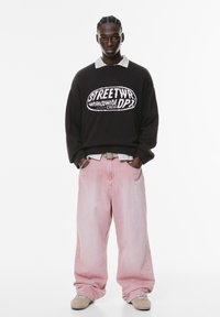 Sort oversized sweatshirt med hvid grafisk tekst, lagdelt over en ternet skjorte, parret med pink vide bukser og beige sneakers.