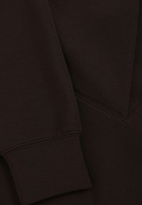 Tejido de sudadera marrón oscuro con una textura suave, que presenta un detalle de costura diagonal y puños acanalados. Diseño simple y minimalista.