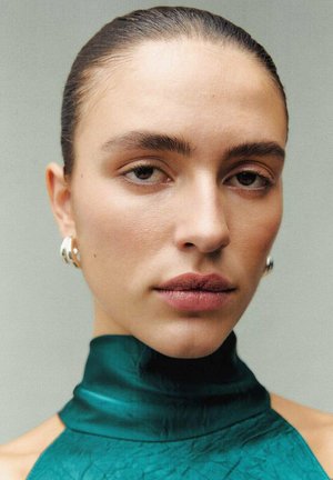 Gros plan sur un visage avec une peau lisse, des sourcils foncés et des lèvres nues. Porte des boucles d'oreilles en argent et un vêtement turquoise à col haut avec une finition texturée.