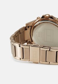 Montre en rose gold métallique avec un bracelet à maillons, dotée d'une surface texturée et d'un logo gravé sur le fermoir. Arrière en verre lisse visible.