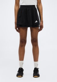 Άτομο που φοράει μαύρη αθλητική φούστα με λευκές ρίγες, λευκές κάλτσες μέχρι τη μέση της γάμπας και μαύρα αθλητικά παπούτσια, στέκεται σε απλό φόντο.