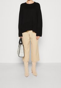 Svart stickad tröja med breda ärmar, beige cropped byxor och beiga klackboots. Håller en vit och svart mönstrad handväska.