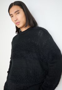 Black knitted sweater with various textures, including smooth and fluffy sections. Crew neckline and relaxed fit, with subtle patterns throughout.