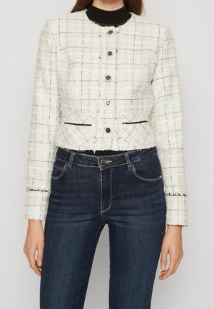 Kvinde iført en hvid tekstureret cropped jakke med sorte gitterlinjer, sølvknapper og mørkeblå denimjeans.