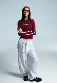 Bordeauxfarbenes Langarmoberteil mit weißen Streifen und Logo; graue lockere Sweatpants; weißer gebundener Pullover; schwarze Schuhe; strukturierter Hintergrund.