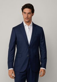 Veste de costume bleu marine en tissu texturé, deux poches avant, fermeture à un bouton, accompagnée d'une chemise blanche.