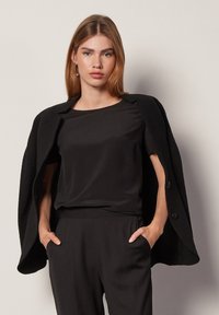 Svart texturerad kappa med oversized siluett, bärs öppen över en slät svart topp och byxor. Utseendet kännetecknas av enkla linjer och knappdetaljer.