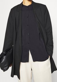 Sort blazer med notchede revers, lagd ovenpå en mørk knap skjorte. Cremefarvede wide-leg bukser og en sort clutch taske fuldender outfittet.