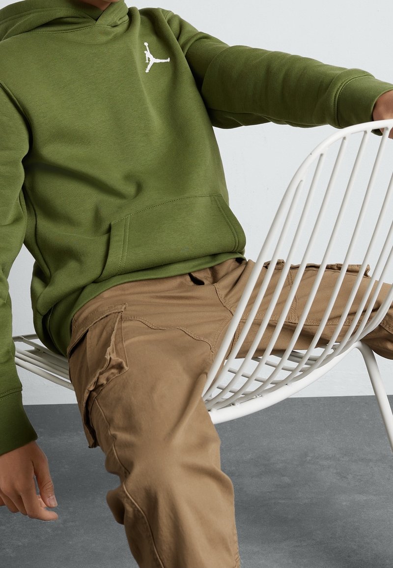 En person klädd i grön huvtröja och beige cargobyxor sitter på en vit trådstol mot en grå golv och vit vägg.