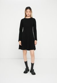 Vestido negro de manga larga con un corsé ajustado, falda acampanada y dobladillo sin terminar. Modelo vistiendo zapatos negros y calcetines gris oscuro.