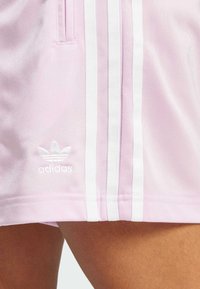 Roza sporta šortus ar trim baltām svītrām gar malām un izšūtu baltu Adidas logo apakšējā kreisajā stūrī. Gluda tekstūra.
