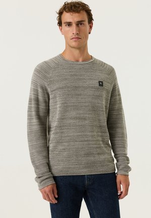 Man with short curly hair wears a gray textured long-sleeve sweater and dark blue jeans, standing against a plain light background.