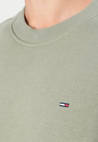 Olijfgroene sweatshirt met geribbelde kraag, voorzien van een klein geborduurd logo in rood, wit en blauw op de borst. Gladde textuur.