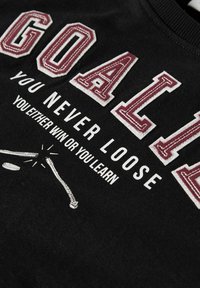 Juodas medvilnės švarkas su ryškiai siuvinėtais baltais ir burgundiškais rašmenimis; užrašas skaito "SARGO, NIEKADA NEPRALAIMI." Piešinys su ledo ritulio lazda ir kamuoliuku.