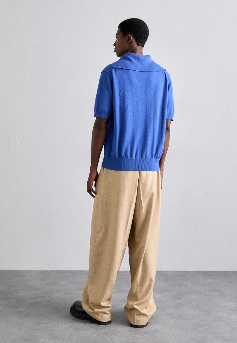 Blauw gebreid polo shirt met korte mouwen, geribbelde zoom, gecombineerd met lichtbeige wijde broek en zwarte schoenen. Glad textuur.