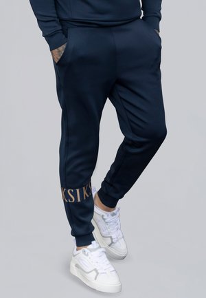 Marinefarbene Jogger-Hosen aus weichem Material mit schlanker Passform, ausgestattet mit Seitentaschen und einem goldenen Logo-Akzent am Saum. Kombiniert mit weißen Sneakers.