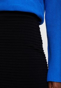 Modrý pletený svetr s ribovanou texturou, zkombinovaný s černou ribovanou sukní s horizontálními pruhy, která zvýrazňuje přiléhavou siluetu.